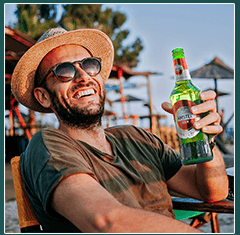 Social media photo: a man in a straw hat holding an Amstel at the beach